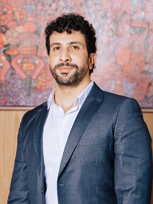 Professional portrait of a man with curly dark hair and a beard, wearing a suit and white shirt, with abstract art in the background.